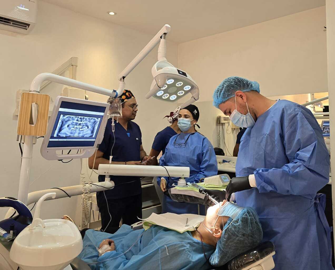 Fotografía en un consultorio dental que muestra a un equipo médico especializado atendiendo a un paciente recostado en el sillón clínico durante un procedimiento bajo sedación consciente, con instrumental clínico y monitores de apoyo visibles.